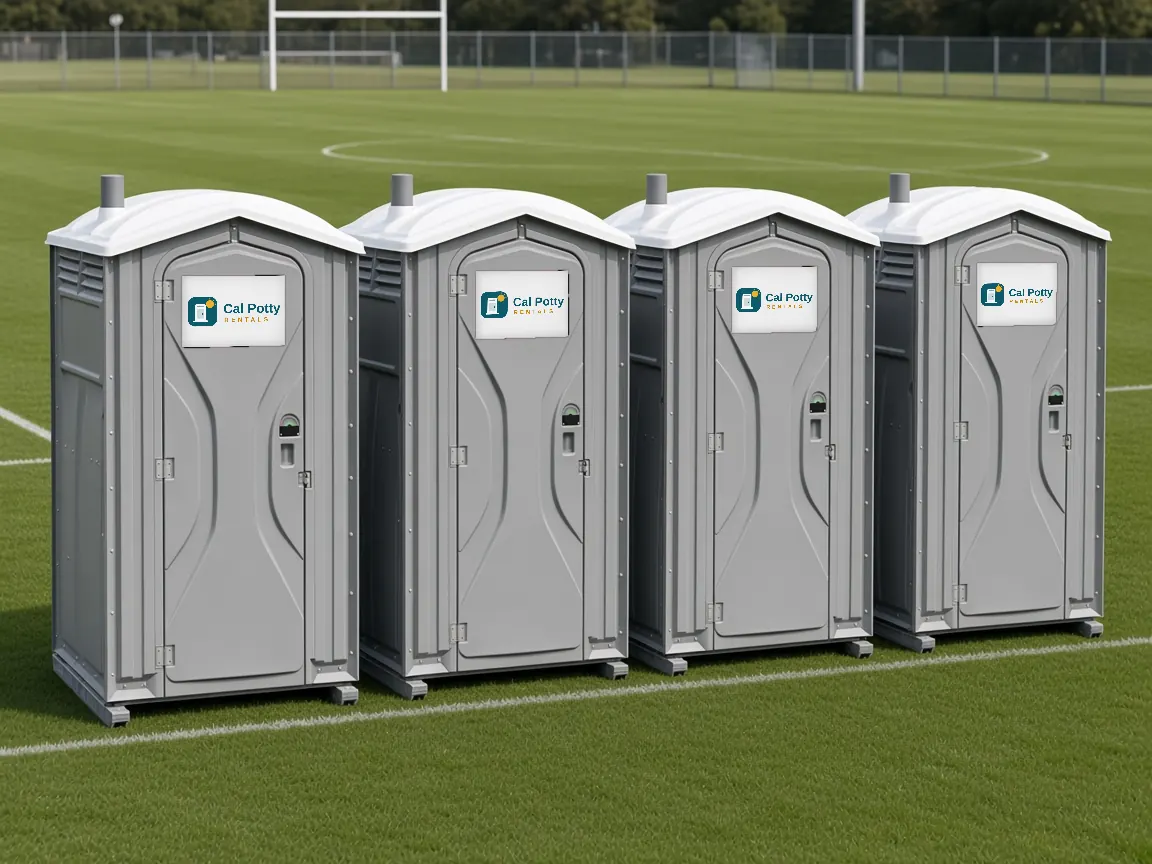 Four grey portable toilets in a row at the edge of a soccer field