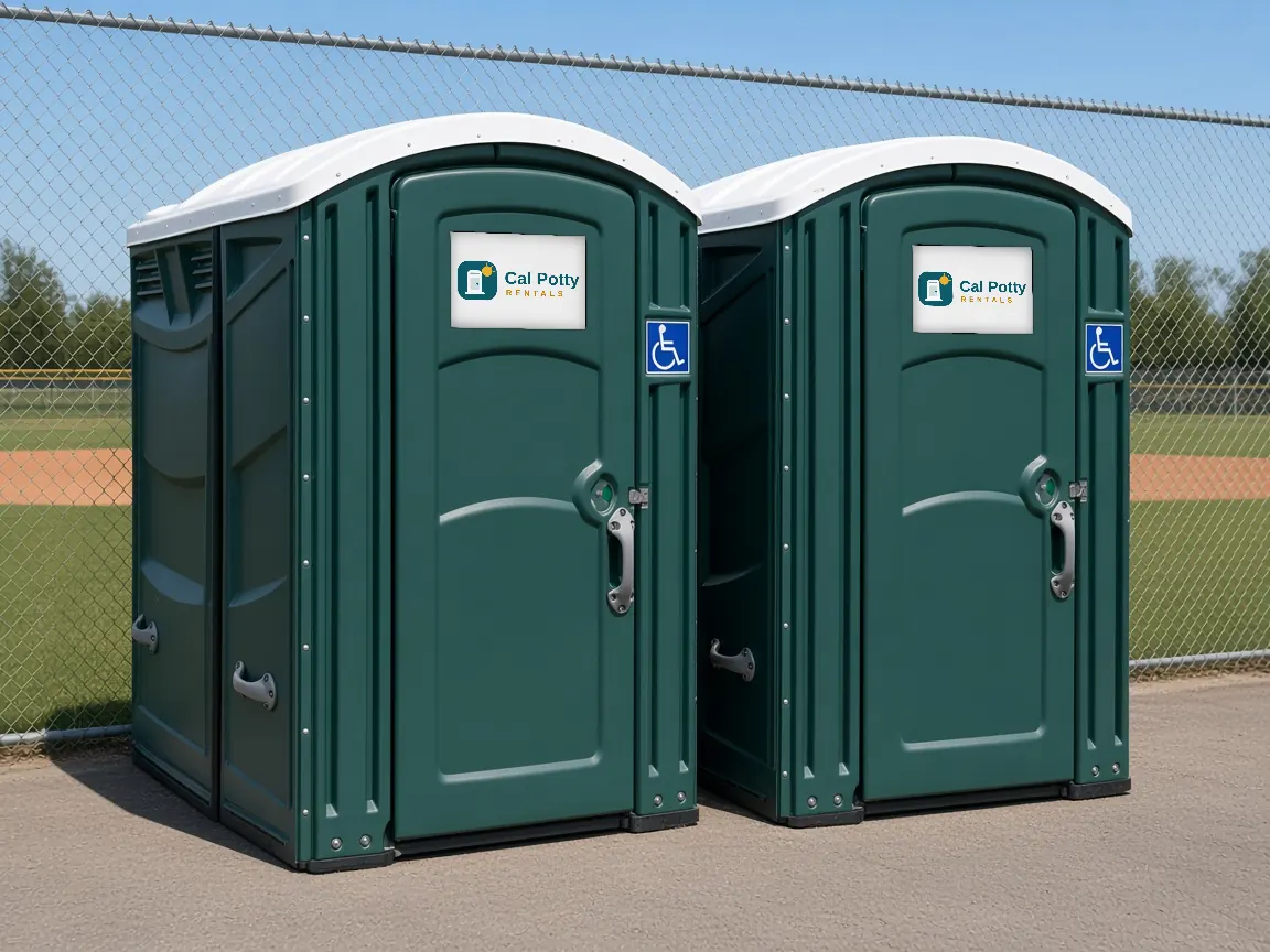 Two teal ADA-accessible portable restrooms with wheelchair signs beside a baseball field fence